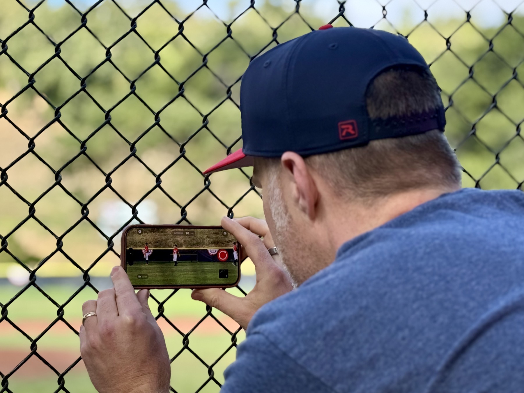 Person recording a baseball game with Sideline Save on an iPhone from behind a chain-link fence