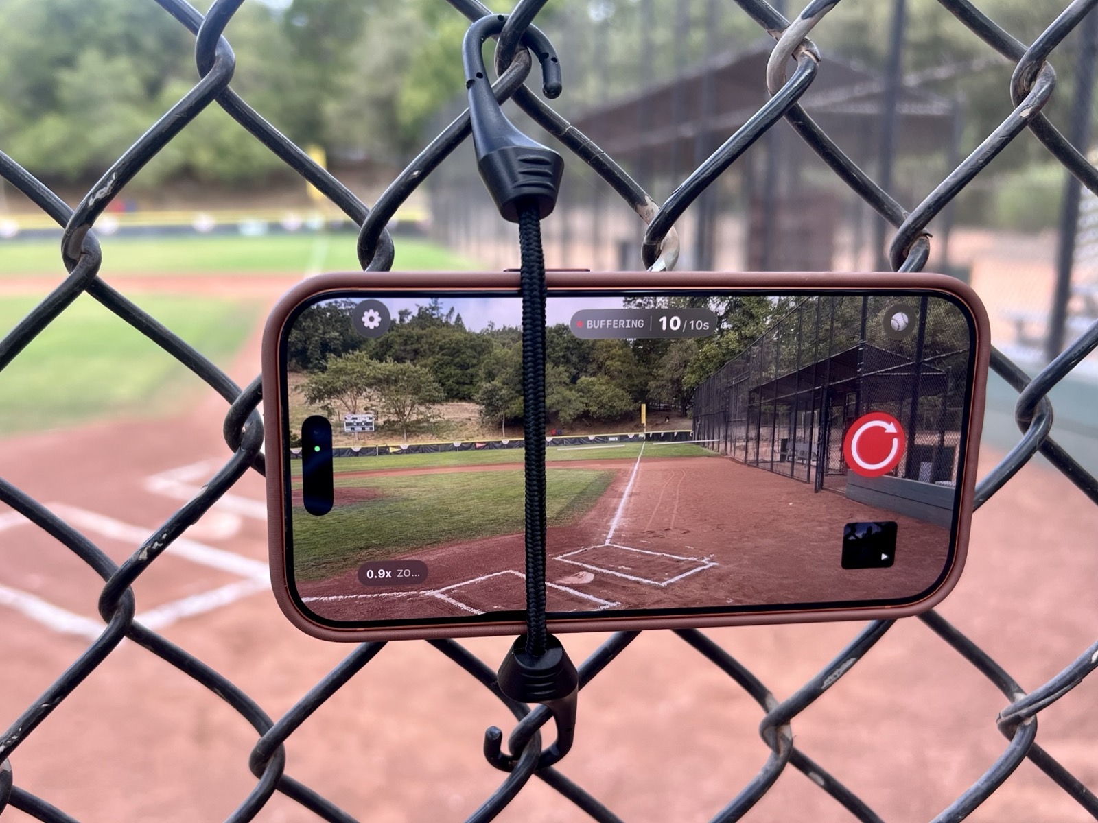 iPhone running Sideline Save attached to a chain-link fence with a short black bungee cord at a baseball field