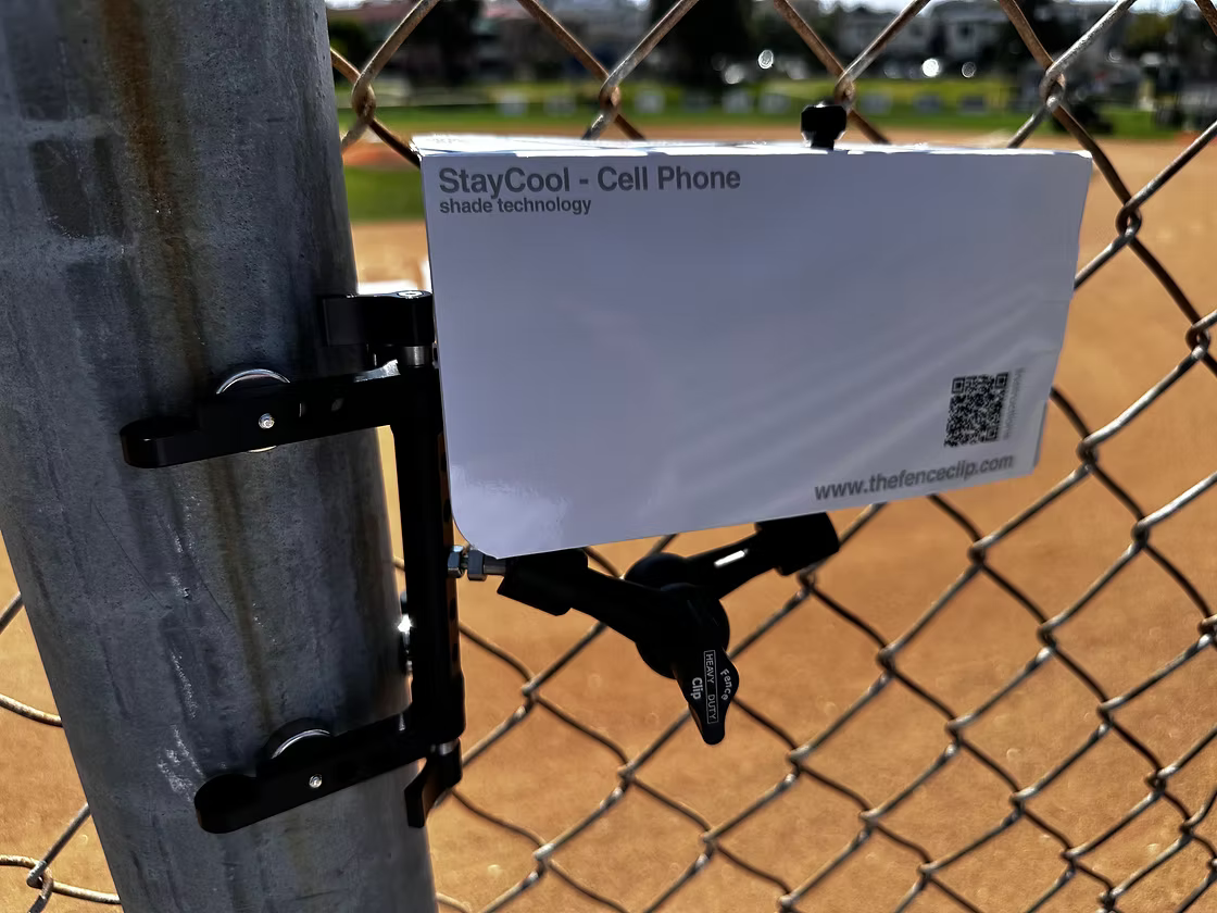 A white FenceClip paper shade mounted over a phone on a chain-link baseball fence